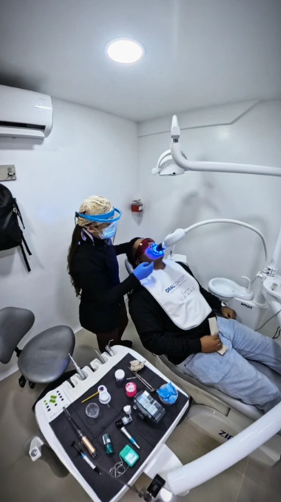 Profesional con mascarilla y guantes atendiendo a un paciente en un sillón dental, junto a un equipo con luz azul.