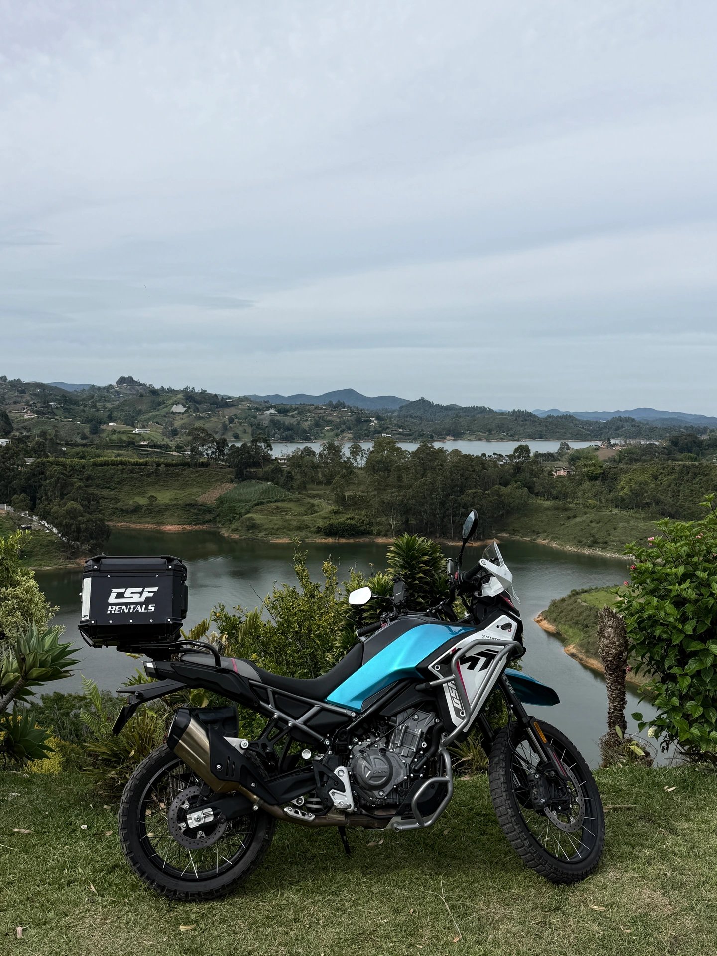 Motocicleta azul y blanca con un baúl de CSF Rentals, estacionada frente a un embalse y montañas.