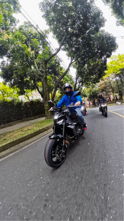 Motociclistas recorren una vía arbolada, con un conductor de chaqueta azul en primer plano.